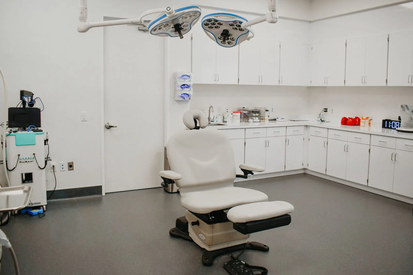 Clinical examination room with surgical lights, patient chair, and white cabinetry stocked with medical supplies